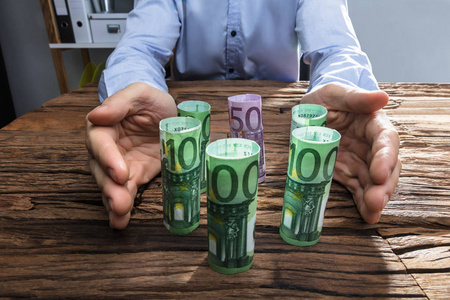 s Hand Protecting The Rolled Up Currency Notes On Wooden Desk