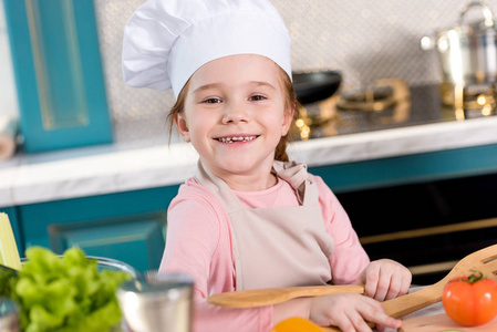 戴着厨师帽和围裙的可爱孩子在厨房做饭时对着镜头微笑