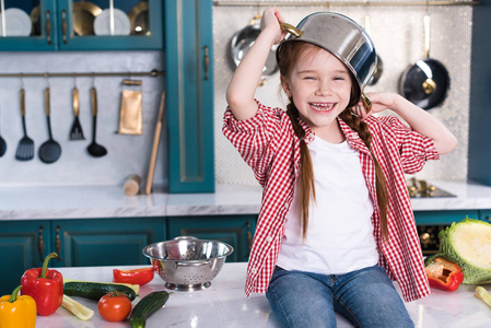 可爱的孩子，头上有平底锅，玩得很开心，坐在厨房的桌子上。