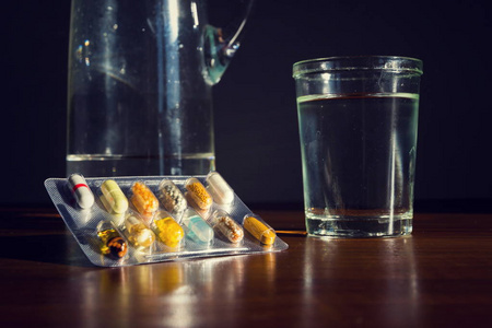 透明胶囊中各种彩色药丸的细节与棕色木桌上的杯水，疾病医学概念
