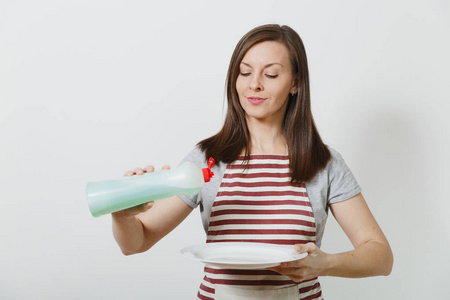 年轻的家庭主妇在条纹围裙隔离白色背景。女管家拿着瓶子用干净的液体洗碗, 白色的空圆盘子。广告复制空间瓶