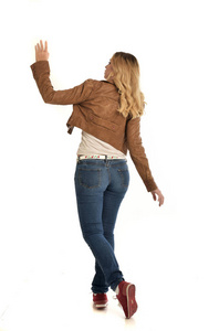 穿棕色夹克和牛仔裤的金发女孩的全长肖像。 站在白色背景上的姿势。
