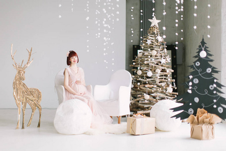 年轻的孕妇坐在白色的椅子上，在冬天的背景上，圣诞树装饰着