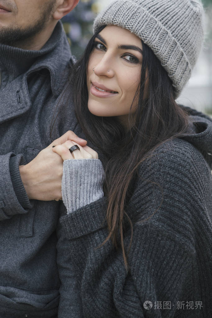 夫妻俩穿着休闲的灰色衣服,拥抱在一起. 天气寒冷,背景上有圣诞灯