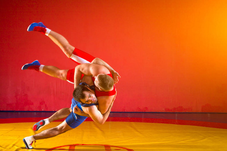 两个穿着蓝色和红色摔跤紧身衣的年轻人正在体育馆里摔跤，在黄色的摔跤地毯上做一个复杂的摔跤