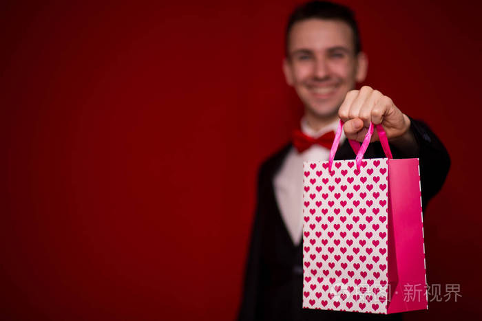 年轻的微笑男人穿西装，拿着红色背景的爱情礼品袋，重点放在礼品袋上。 情人节作文