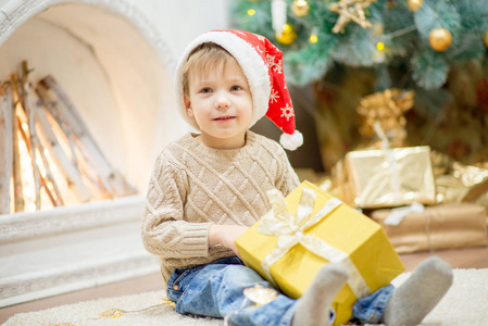 一个戴红帽的男孩坐在圣诞树旁, 带着礼物