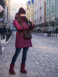 穿着紫色冬季大衣, 帽子和围巾的漂亮黑发女孩, 在冬天走欧洲街, 在她的智能手机上拍照, 使自拍