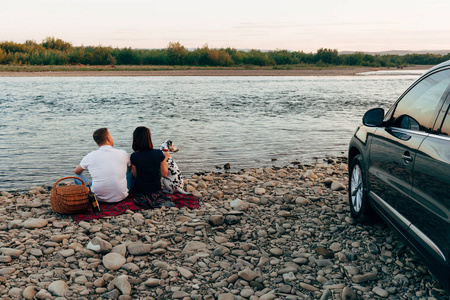 幸福的年轻夫妇与狗在 roadtrip 的肖像。男人和女人坐在格子上户外野餐概念