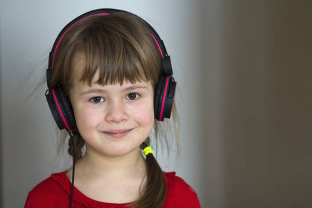 照片的快乐漂亮的儿童小女孩与大耳机在家里。快乐的儿童女孩听音乐在灰色模糊背景。首页技术与音乐理念