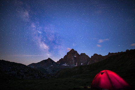 在高空上阿尔卑斯山野营下满天星斗的天空和银河系。在背景前景和雄伟的山峰的照明的帐篷。冒险和探索在夏天