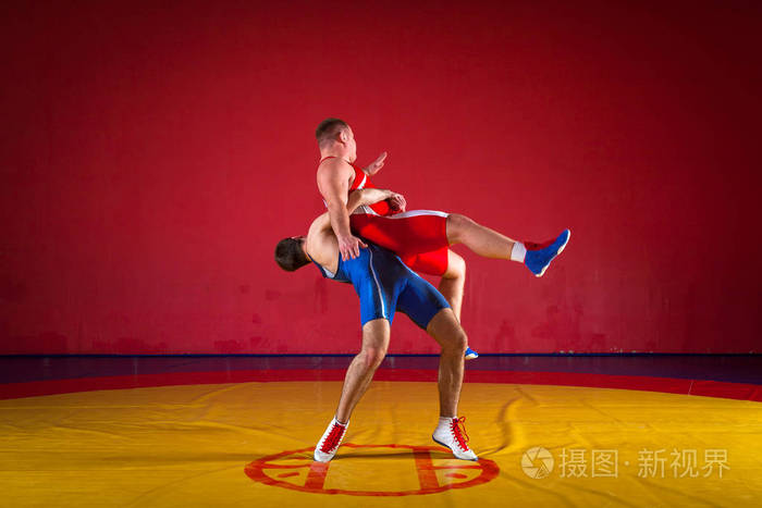 两位身穿红蓝制服的罗马摔跤手在体育馆的黄色摔跤地毯上摔跤