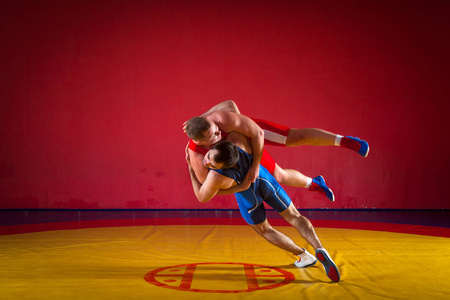 两个穿着蓝色和红色摔跤紧身衣的年轻人正在体育馆里摔跤，在黄色的摔跤地毯上做一个复杂的摔跤