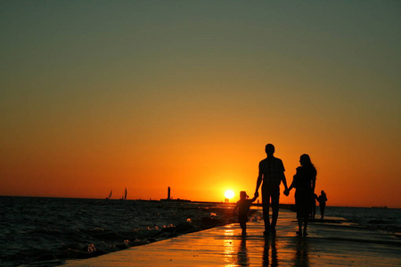 一家三口在堤坝上行走的剪影,双手握着明亮的夕阳,背景是晴朗的天空.