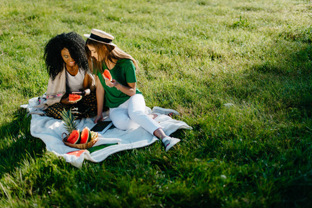 野餐主题。两个诱人迷人的非洲和高加索女孩朋友聊天和浏览通过手机, 而吃西瓜