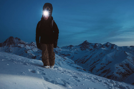 勇敢的夜间探险家爬上高山。 戴前照灯背包和滑雪服。 滑雪者背着一块滑雪板在雪山上行走。 内陆国家