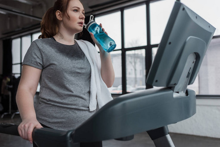 身材曲线美的女孩在健身房跑步机上跑步时，从水瓶里喝水
