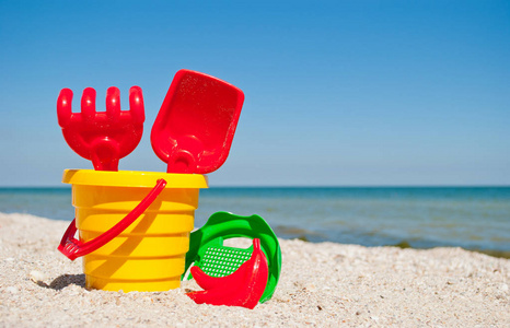 黄色的婴儿桶，有红色的手柄，塑料的红色铲子，耙和绿色的筛子，一个红色的沙子形式，香蕉，蓝色的大海和天空，黄色的沙滩，海岸，贝壳，