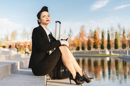 穿着制服的年轻女空姐坐在公园里带着手提箱, 等待着她的飞行。