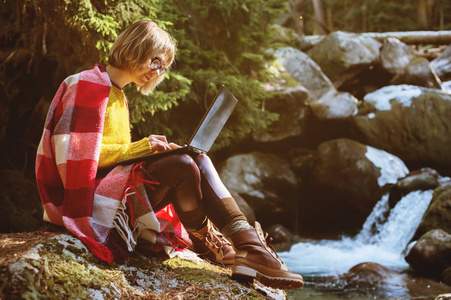 一个色调的肖像, 一个微笑的自由职业者时髦的女孩戴着眼镜, 穿着毯子与膝上型电脑跪在一座针叶林的岩石旁边的暴风雨山河在温暖