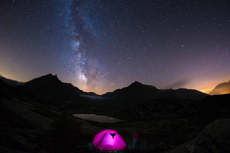 在高空上阿尔卑斯山野营下满天星斗的天空和银河系。在背景前景和雄伟的山峰的照明的帐篷。冒险和探索在夏天