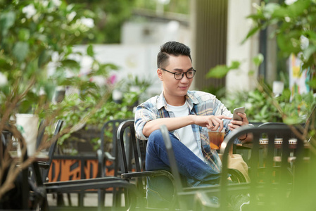 戴眼镜的英俊亚洲男人，端着冰茶坐在户外，在智能手机上使用应用程序