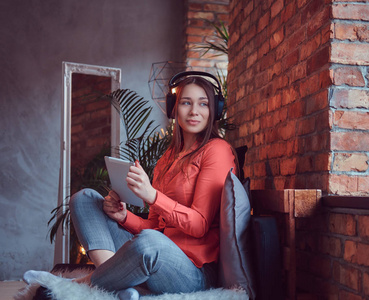 一个迷人的微笑黑发女孩穿着休闲服装，使用平板电脑，同时通过良好的耳机听令人兴奋的音乐。 在有阁楼内部的房间里放松。