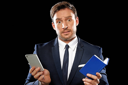 英俊的聪明商人穿着一套经典的办公室西装和领带，手里拿着一本笔记本和一部手机，显示出一项艰苦的工作。 孤立在黑色背景上