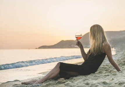 年轻的金发女人在日落时在海边的海滩上享受一杯玫瑰酒。埃及艳后海滩，阿拉尼亚，地中海地区，土耳其。