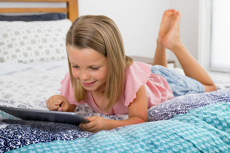 美丽的金发6或7岁的女孩躺在床上微笑快乐使用互联网上的数字平板电脑垫观看和乐趣