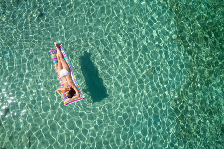 穿着鲜艳比基尼的年轻女人的俯视图是在透明的蓝色海洋中游泳。躺在水面上漂浮的苗条女人的空中景色