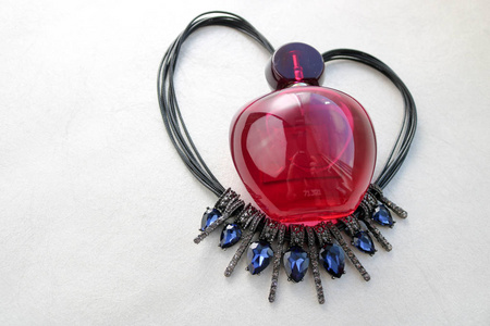 红色，美丽的玻璃，透明的女性香水瓶，躺在黑色的绳子上，形状是一颗用拖着的蓝色钻石装饰的心，在浅白色的背景上放置一个简单的文字。