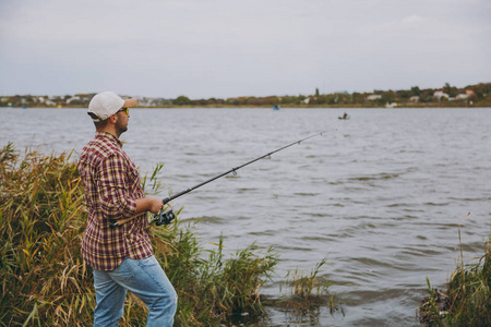 侧面查看年轻的刮胡子男子与钓鱼竿在格子衫, 帽子和太阳镜在湖上的钓鱼竿在灌木和芦苇附近的岸边。生活方式, 休闲, 渔民休闲概念