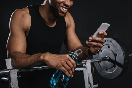 微笑的年轻非洲裔美国运动员拿着一瓶水，用智能手机拍摄的镜头