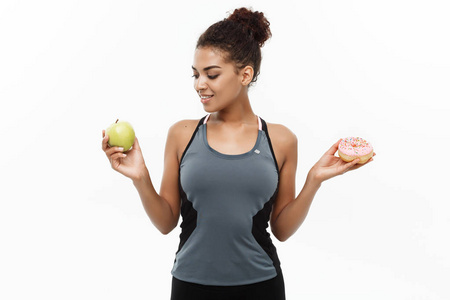 健康和饮食概念美丽的运动的非洲裔美国人在甜甜圈和绿色苹果之间做出决定。在白色背景上被隔离