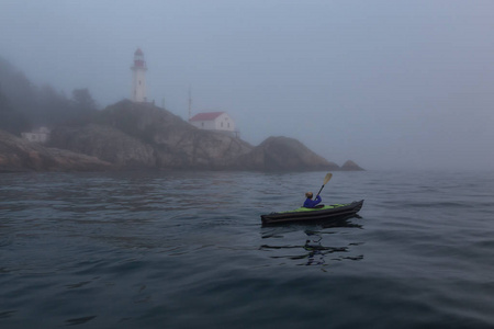 冒险的女人是在灯塔附近的海上皮划艇，在一个充满活力和雾的冬季日落。 在加拿大温哥华西马蹄湾拍摄。 概念冒险假期探索