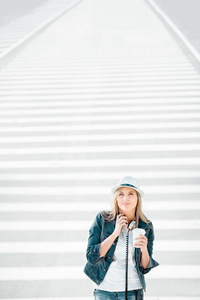 美丽的年轻女子戴着帽子和牛仔裤夹克，带着一个外卖咖啡杯站在路上，斑马线喝着咖啡，在道路背景下摆姿势。