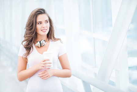 快乐的年轻女子戴着音乐耳机，站在桥上，拿着一个拿走的咖啡杯，在城市背景下眺望。