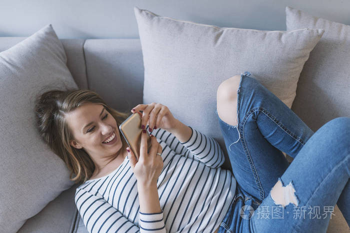 女孩在智能手机上阅读信息。 幸福的女人在沙发上用手机。 幸福的女人在家里在她明亮的客厅里读短信。 放松的年轻女子在沙发上发短信