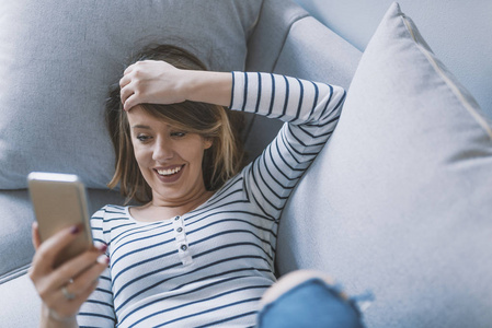 女孩在智能手机上阅读信息。幸福的女人在沙发上用手机。快乐的女人在她明亮的客厅里看短信。沙发上轻松的年轻女性短信