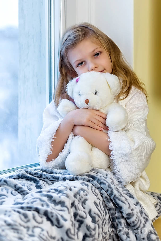 可爱的小女孩抱着泰迪熊.一个可爱的婴儿在房间里坐在窗口在冬天