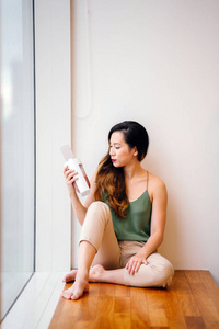 年轻的亚洲中国女人早上喝一杯透明不倒翁的健康混合奶昔。 她坐在她公寓的窗户旁，阳光洒了进来。 她穿得很舒服。