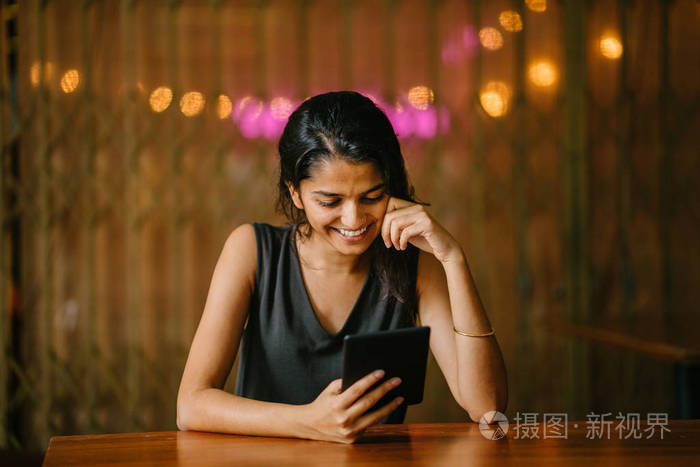 坦率的肖像，一个有吸引力和年轻的印度亚洲职业妇女阅读她的平板电脑或电子阅读器。 她在温暖舒适的咖啡馆或同事空间。 当她读她的平板