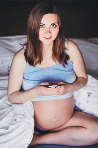 年轻迷人的孕妇坐在床上, 手里拿着手机。美丽的孕妇在卷起罐顶与裸露的腹部坐在床上, 拿着手机