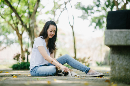 一位年轻的泛亚女人和她年轻的石子小狗坐在公园里的肖像。 她在微笑，他们都在享受温暖的一天。