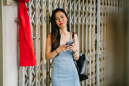 一位年轻的上镜和迷人的中国亚洲女人的肖像，她带着智能手机，而她在亚洲的一条街上等待她的约会。 她微笑着，看上去很高兴。