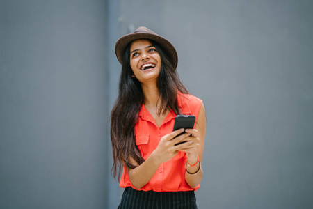 一位年轻时尚的北印度人亚洲女人的肖像。 她肤色浅，穿着一件橙色衬衫，黑色裤子和一顶联邦帽。 她微笑着使用她的智能手机。