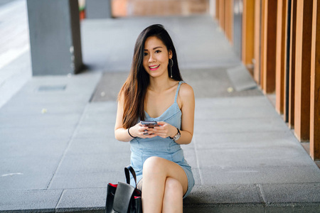 一位年轻的上镜和迷人的中国亚洲女人的肖像，她带着智能手机，而她在亚洲的一条街上等待她的约会。 她微笑着，看上去很高兴。
