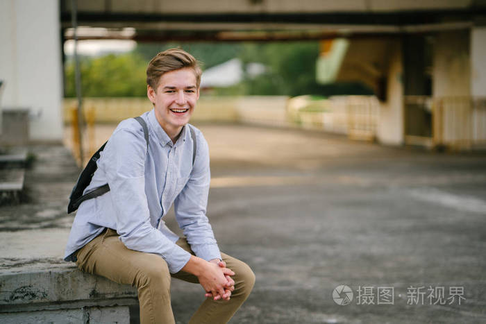 一个年轻而有吸引力的白种人游客或学生坐在亚洲的一个混凝土停车场日出或日落时微笑的肖像。 他在微笑，很放松。