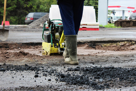 一名道路施工工人用一个紧凑的振动板压实土壤，然后在道路的一个有问题的沼泽部分沥青。俄罗斯，进入加契纳市..